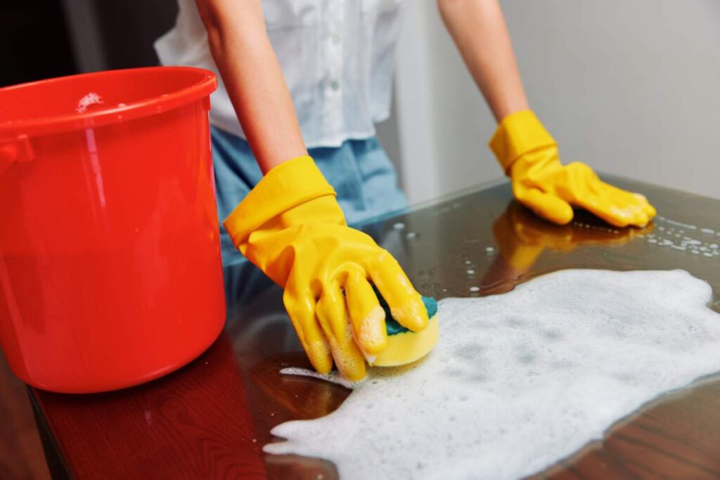 Woman cleaning table with soapy water and red bucket wearing yellow rubber gloves.